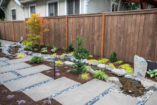 Backyard landscaping with large concrete stepping pavers, mulched planting beds, and a small stream beside a wooden fence in the San Fernando Valley